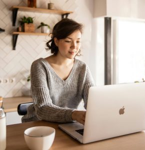 Young female in gray sweater sitting at wooden desk with laptop and bottle of milk near white bowl while browsing internet on laptop during free time at home